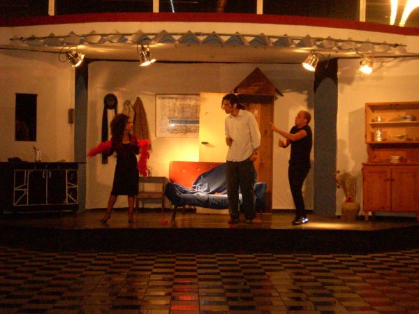 Tre persone recitano su un palco con arredi casalinghi.