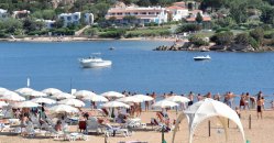 Spiaggia affollata con lettini e ombrelloni bianchi, mare e barche in lontananza.