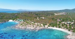 Costa rocciosa con acqua turchese, vegetazione bassa e colline sullo sfondo.