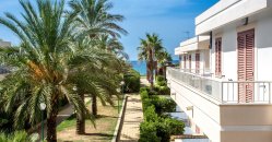 Palazzina bianca con palme e vista sul mare in lontananza.