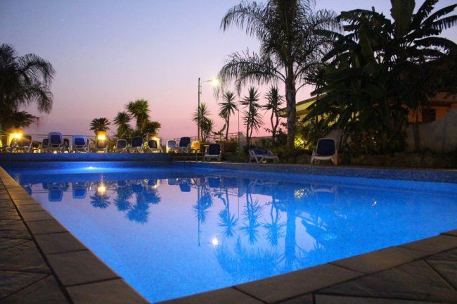 Piscina illuminata al tramonto con palme e cielo viola.