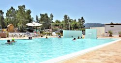 Piscina all'aperto con alcune persone che nuotano, ombrelloni e sedie a sdraio sullo sfondo.