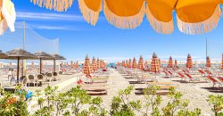 Spiaggia soleggiata con ombrelloni e lettini, vista da sotto un grande ombrellone arancione.