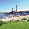 Spiaggia affollata con ombrelloni e edificio con ciminiere.
