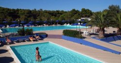 Una piscina con bagnanti, circondata da lettini e ombrelloni blu, con alberi e colline sullo sfondo.