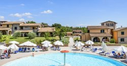 Piscina circondata da ombrelloni e lettini, con edifici e giardino sullo sfondo.