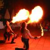 Due artisti di strada sputano fuoco davanti a uno spettacolo serale con pubblico seduto.