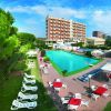 Piscina con sdraio e tavoli rossi, circondata da giardino e grandi edifici sullo sfondo.
