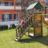 Parco giochi con scivolo e casetta in legno, bambini che giocano. Sfondo con edificio arancione.