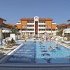 Piscina con vari nuotatori e sdraio intorno. Edifici arancioni e cielo blu sullo sfondo.
