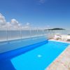 Una piscina azzurra su una terrazza con lettini bianchi e un cielo blu con alcune nuvole.
