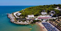 Costa rocciosa con spiaggia attrezzata e case bianche tra la vegetazione; mare blu sullo sfondo.