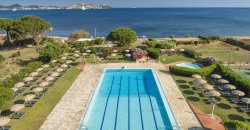 Piscina rettangolare vicino al mare, circondata da ombrelloni e lettini.