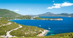 Una baia con mare blu e stradina costiera, montagne verdi in lontananza.