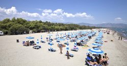 Spiaggia con numerosi ombrelloni blu e lettini, circondata da alberi e montagne in lontananza.
