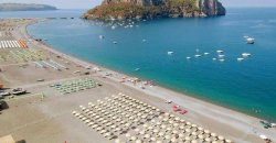 Spiaggia con file di ombrelloni e barche ancorate in mare, isola collinare sullo sfondo.