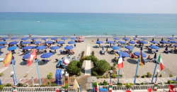 Spiaggia con ombrelloni blu e bandiere nazionali, mare calmo sullo sfondo.