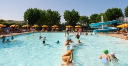 Una piscina affollata con scivolo, ombrelloni gialli, alberi sullo sfondo.