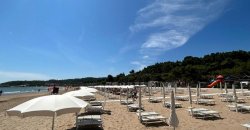 Spiaggia sabbiosa con lettini bianchi e colline verdi in lontananza.