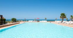 Piscina con vista sul mare, lettini bianchi e palme lungo il bordo.