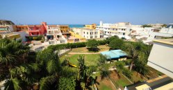 Vista su un complesso di edifici colorati vicino al mare con un prato e palme in primo piano.