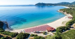 Spiaggia con ombrelloni rossi su sabbia bianca, mare limpido e montagne in lontananza.